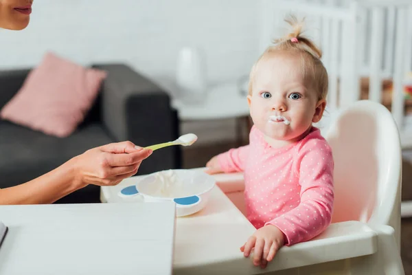 Selektiver Fokus des Säuglings mit unordentlichem Mund, der in die Kamera neben der Mutter mit einem Löffel Babynahrung blickt — Stockfoto
