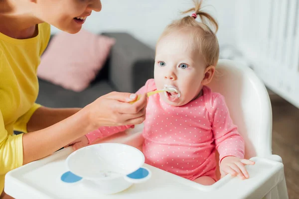 Concentration sélective de la femme nourrissant bébé fille sur chaise haute à la maison — Photo de stock