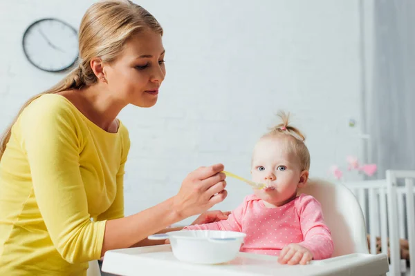 Женщина держит ложку детского питания во время кормления дочери на высоком стульчике дома — стоковое фото