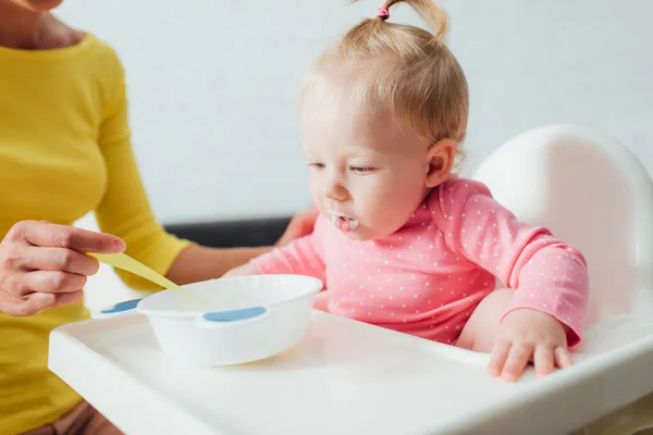 Selective focus of infant girl sitting in highchair near mother with spoon and bowl of baby food — Stock Photo