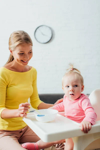 Concentration sélective de la mère regardant bébé fille près du bol et cuillère sur la chaise d'alimentation — Photo de stock