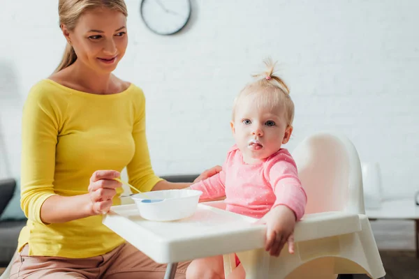 Concentration sélective de bébé fille regardant la caméra près de la mère avec cuillère et bol de nourriture pour bébé à la maison — Photo de stock