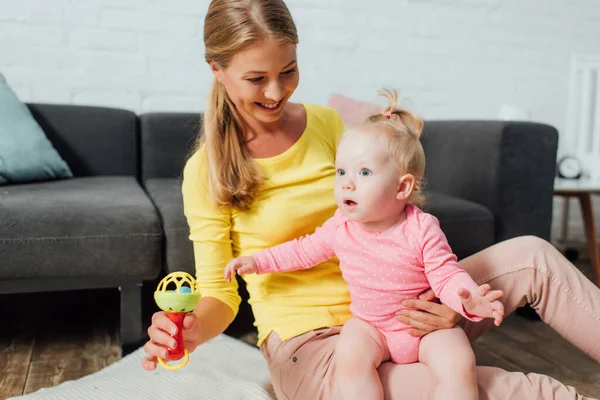 Mother holding rattle and excited baby daughter on floor at home — Stock Photo