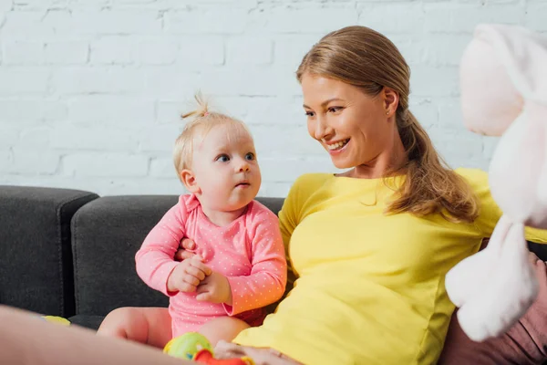 Selektiver Fokus der Mutter, die ihre kleine Tochter in der Nähe von Plüschtier auf der Couch betrachtet — Stockfoto