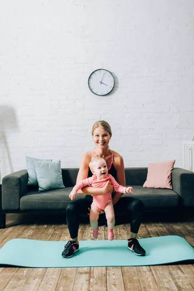 Deportiva sosteniendo a su hija bebé mientras entrena en la colchoneta de fitness en casa - foto de stock