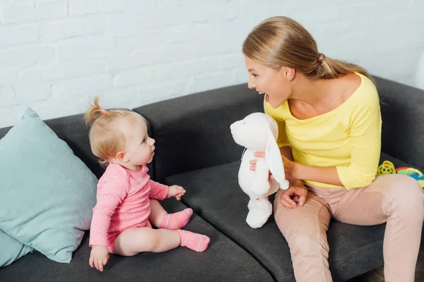 Mother holding soft toy near infant daughter on sofa — Stock Photo