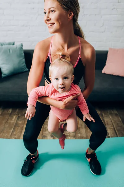 Mother in sportswear holding baby girl while training on fitness mat at home — Stock Photo