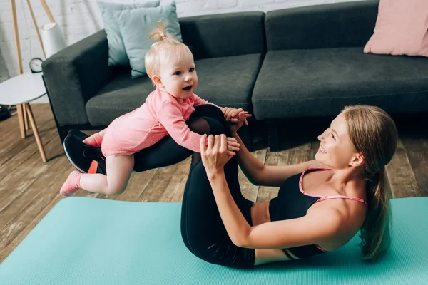 Женщина с маленькой дочкой на руках во время тренировки на фитнес-коврике дома — стоковое фото
