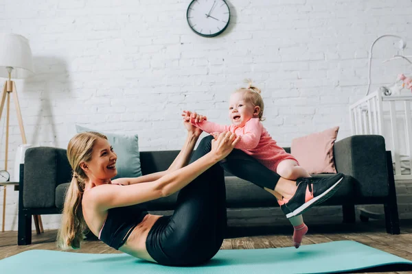 Femme en tenue de sport tenant sa fille tout en faisant de l'exercice sur un tapis de fitness dans le salon — Photo de stock