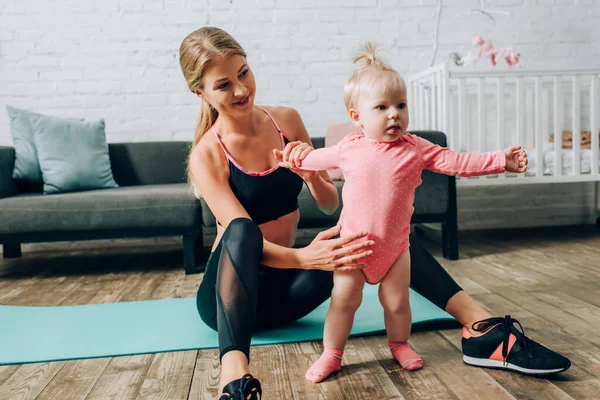Deportiva sosteniendo hija bebé mientras está sentado en la alfombra de fitness en la sala de estar - foto de stock