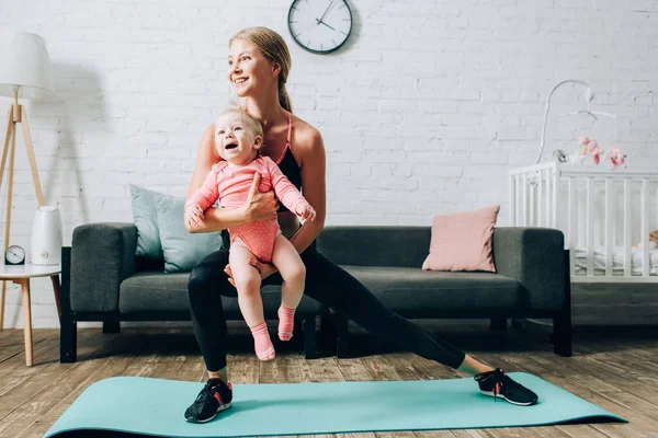 Mujer entrenando en colchoneta de fitness y sosteniendo a su hija en casa - foto de stock
