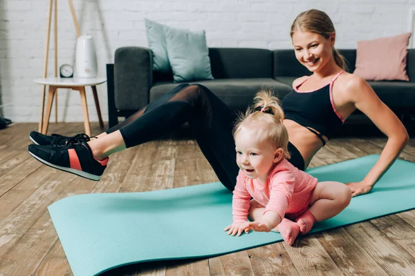 Concentration sélective de bébé fille assis sur tapis de fitness pendant l'entraînement de la mère à la maison — Photo de stock