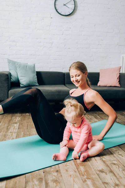 Focus selettivo della bambina seduta sul tappeto fitness mentre la madre si allena a casa — Foto stock