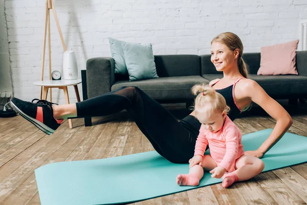 Foco seletivo do treinamento da mulher no tapete de fitness perto da filha do bebê na sala de estar — Fotografia de Stock