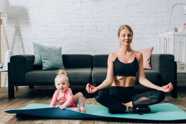 Mulher sentada em pose de ioga no tapete de fitness perto de bebê menina — Fotografia de Stock
