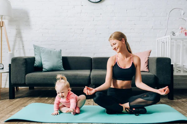 Femme regardant bébé fille tout en méditant sur tapis de fitness à la maison — Photo de stock