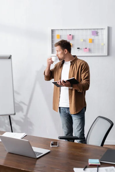 Young businessman in casual clothes drinking coffee and holding notebook while standing in office — Stock Photo