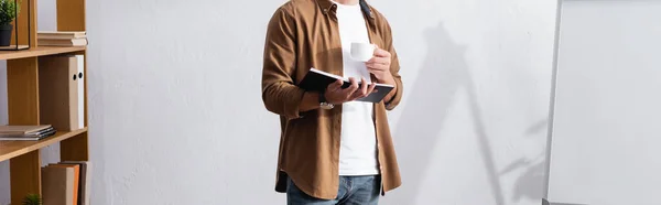 Vista recortada de hombre de negocios en ropa casual sosteniendo portátil y taza de café en la oficina, concepto panorámico - foto de stock