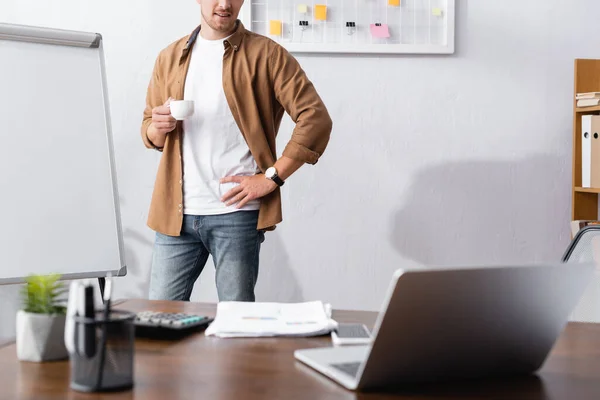 Abgeschnittene Ansicht eines Geschäftsmannes in lässiger Kleidung, der eine Kaffeetasse hält, während er mit der Hand auf der Hüfte im Büro steht — Stockfoto