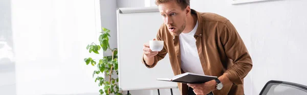 Horizontal image of shocked businessman with coffee cup and notebook in office — Stock Photo