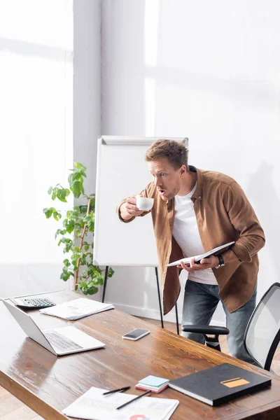 Sorprendido hombre de negocios con la boca abierta mirando a la computadora portátil mientras sostiene el portátil y la taza de café - foto de stock