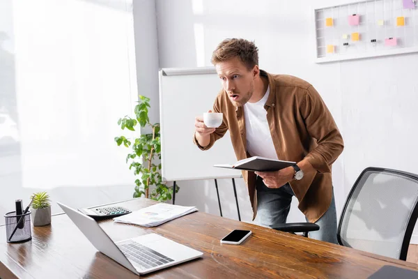 Geschockter Geschäftsmann blickt auf Laptop und hält Kaffeetasse und Notizbuch in der Hand — Stockfoto