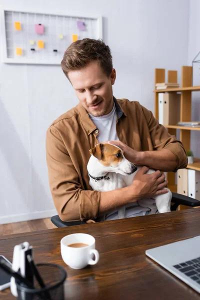Бізнесмен в повсякденному одязі приборкаючи джек-російський тер'єр собака сидить на робочому місці біля чашки кави — стокове фото