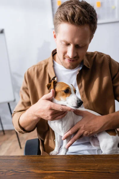 Молодой бизнесмен в повседневной одежде держит Джека Рассела терьера собаки во время сидения на рабочем месте — стоковое фото
