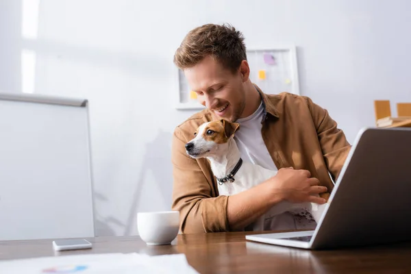 Jovem empresário com jack russell terrier cão sentado no local de trabalho perto de laptop — Fotografia de Stock