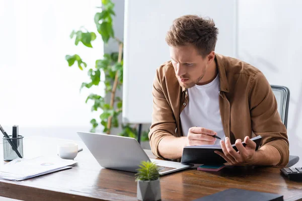 Konzentrierter Geschäftsmann schaut auf Laptop, während er Notizbuch und Stift in der Hand hält — Stockfoto