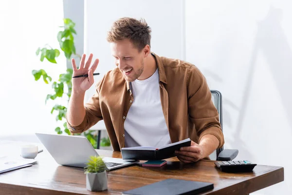 Hombre de negocios saludando la mano durante el chat de vídeo mientras sostiene el cuaderno y la pluma - foto de stock