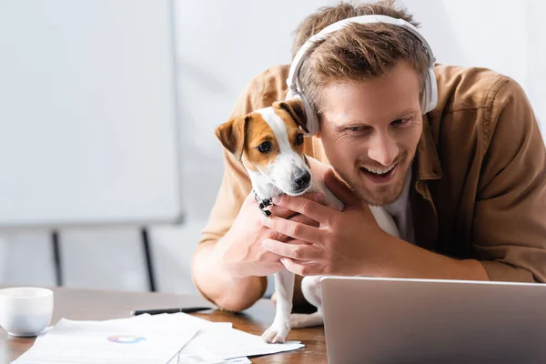 Jungunternehmer mit drahtlosen Kopfhörern kuschelt Jack Russell Terrier Hund am Arbeitsplatz in der Nähe von Laptop — Stockfoto