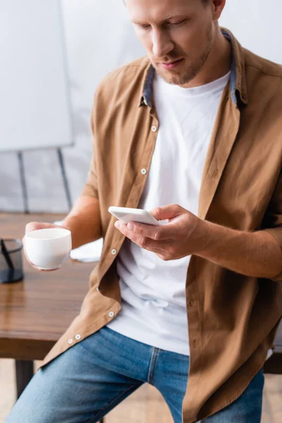 Joven hombre de negocios en ropa casual mensajes de texto en el teléfono inteligente mientras sostiene la taza de café - foto de stock