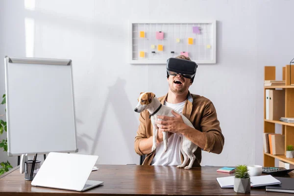 Excitado hombre de negocios en vr auriculares riendo mientras está sentado en el lugar de trabajo con jack russell terrier perro - foto de stock