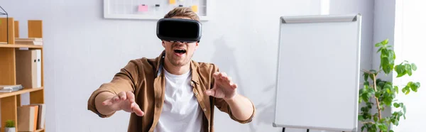 Horizontal image of young businessman with open mouth using vr headset and gesturing at workplace — Stock Photo