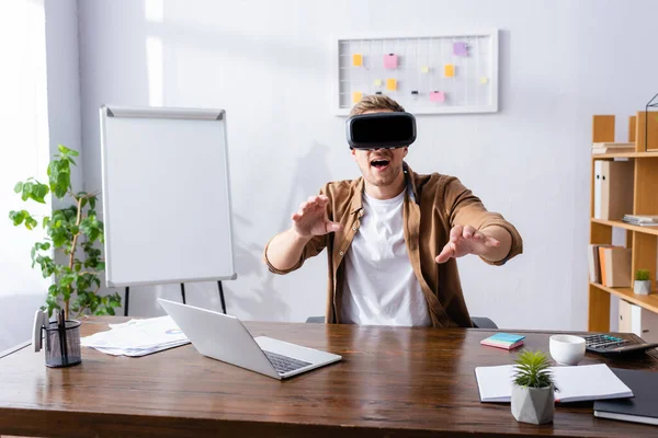 Возбужденный бизнесмен в VR наушники жестом на рабочем месте возле ноутбука — стоковое фото