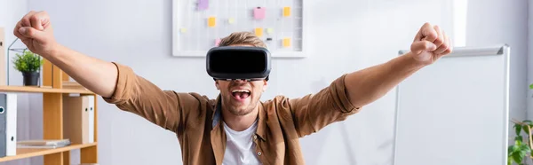 Panoramic shot of excited businessman in vr headset showing winner gesture in office — Stock Photo