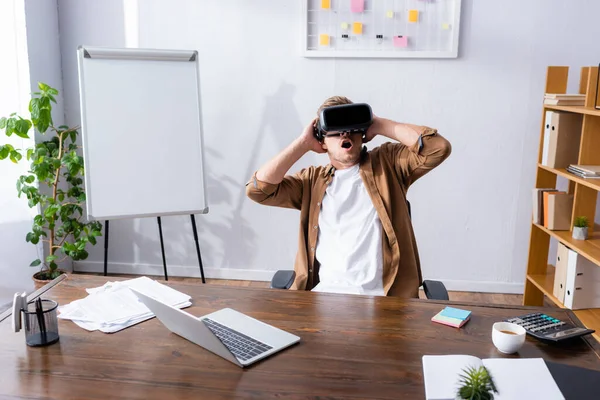 Испуганный бизнесмен с открытым ртом касаясь головы при использовании VR гарнитуры в офисе — стоковое фото