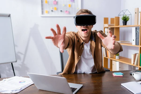 Uomo d'affari eccitato in auricolare vr gesticolare vicino laptop sul posto di lavoro — Foto stock