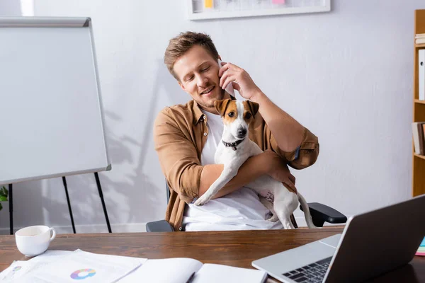 Молодий бізнесмен розмовляє по смартфону, сидячи на робочому місці з джек-рос-тер'єр собакою — стокове фото