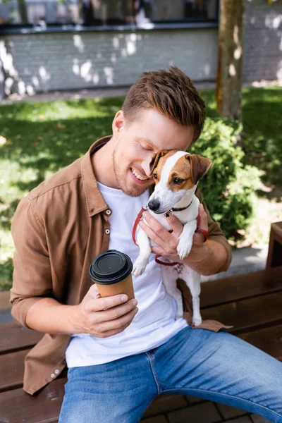 Homme avec les yeux fermés assis sur le banc, câlin Jack Russell Terrier chien et tenant tasse en papier — Photo de stock