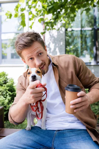 Jeune homme avec Jack Russell terrier chien tenant café pour aller et pointant du doigt tout en étant assis sur le banc — Photo de stock