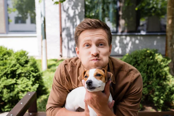 Young man grimacing and puffing out cheeks while holding jack russell terrier dog outdoors — Stock Photo