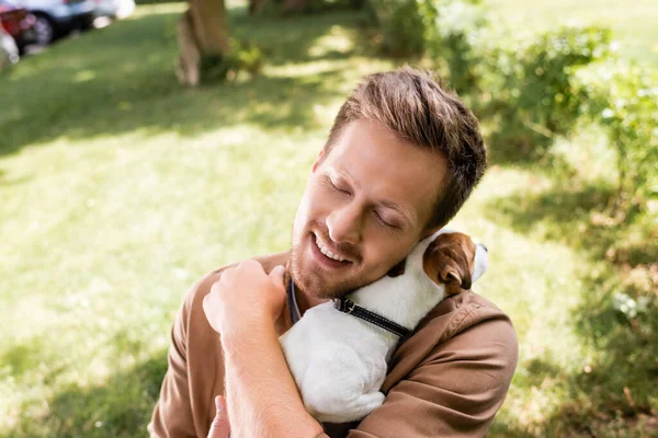 Молодой человек с закрытыми глазами держит и обнимает Джека Рассела терьера собака в парке — стоковое фото