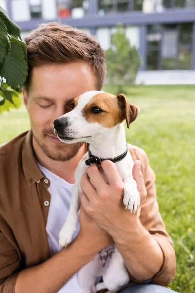 Junger Mann mit geschlossenen Augen hält Jack Russell Terrier Hund mit braunen Flecken auf dem Kopf — Stockfoto