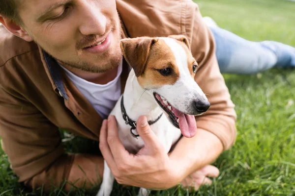 Visão parcial do jovem abraçando Jack Russell terrier cão enquanto relaxa na grama verde — Fotografia de Stock