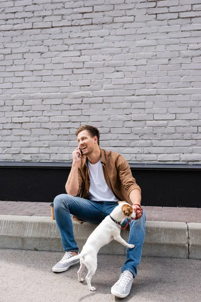 Joven hablando en smartphone cerca de jack russell terrier con correa en la calle urbana - foto de stock