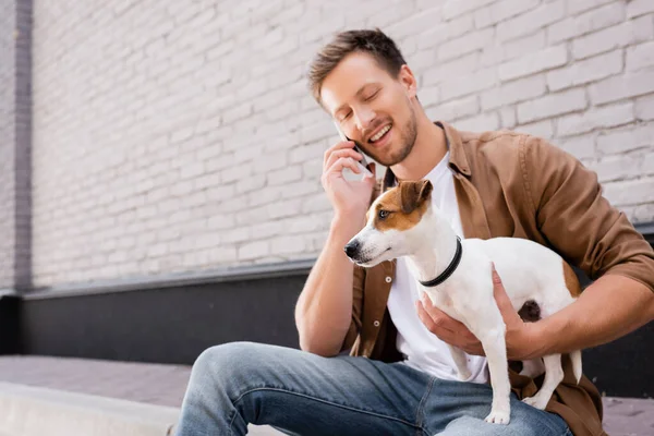 Young man talking on smartphone and hugging jack russell terrier on sidewalk near building — Stock Photo