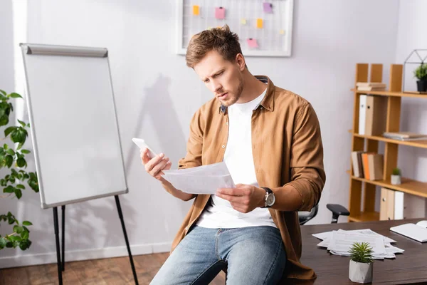 Konzentrierter Geschäftsmann schaut sich Dokumente an, während er sein Smartphone im Büro hält — Stockfoto