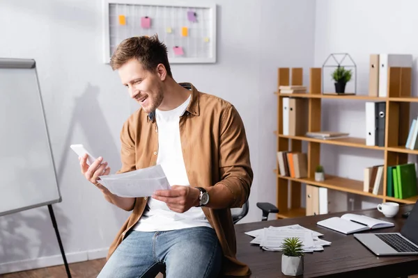 Jeune homme d'affaires utilisant un smartphone et tenant des papiers sur la table au bureau — Photo de stock